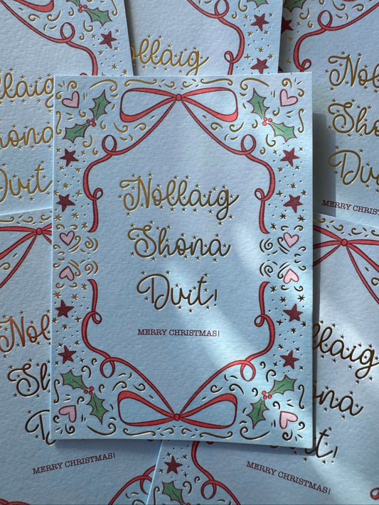 A Christmas greeting card with a white background, decorated with red ribbon bows, green holly leaves, pink hearts, and gold foil lettering that reads 'Nollaig Shona Duit!' and 'MERRY CHRISTMAS!'.