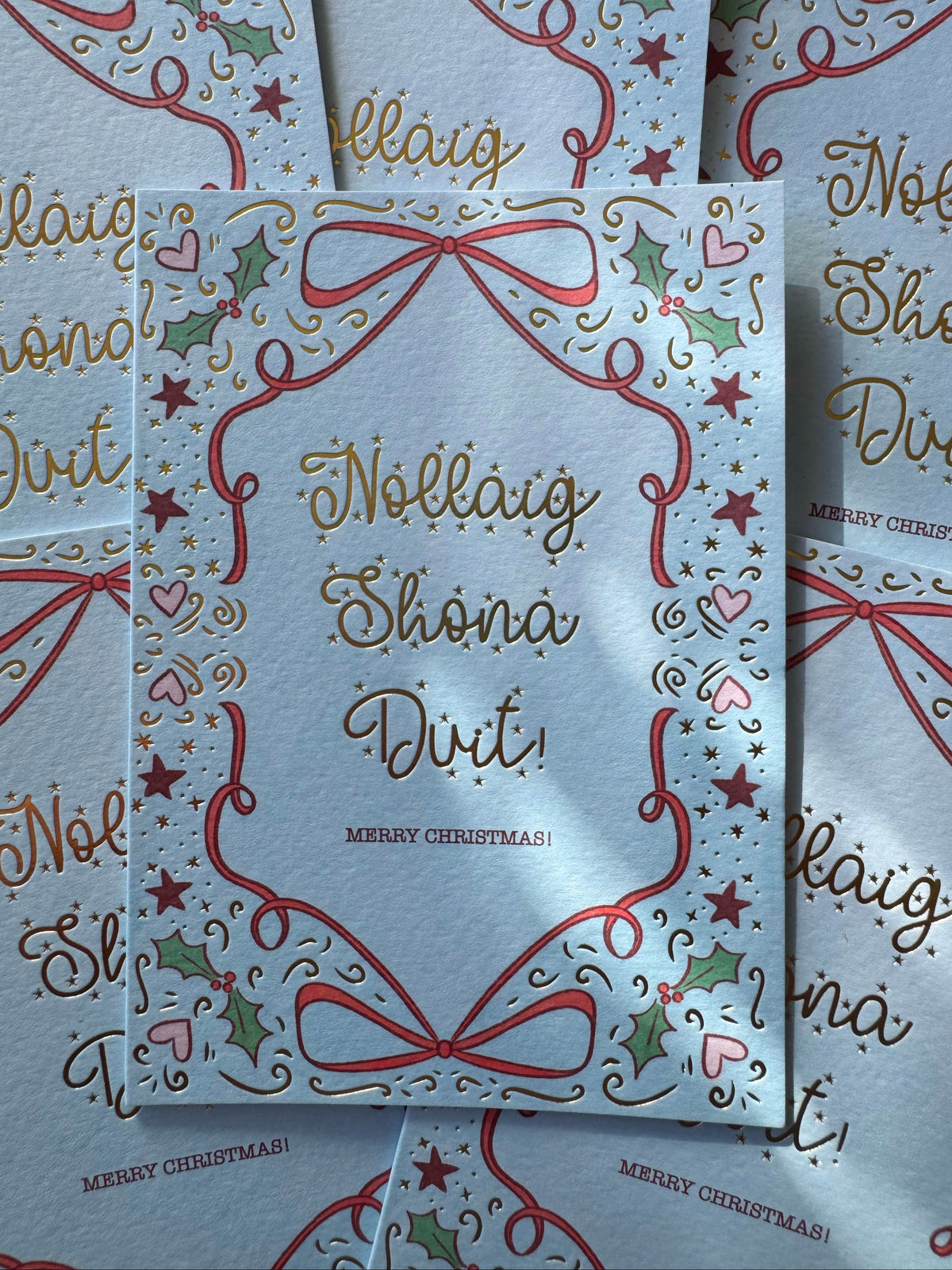 A Christmas greeting card with a white background, decorated with red ribbon bows, green holly leaves, pink hearts, and gold foil lettering that reads 'Nollaig Shona Duit!' and 'MERRY CHRISTMAS!'.