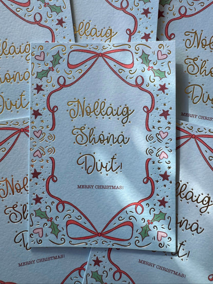 A Christmas greeting card with a white background, decorated with red ribbon bows, green holly leaves, pink hearts, and gold foil lettering that reads 'Nollaig Shona Duit!' and 'MERRY CHRISTMAS!'.
